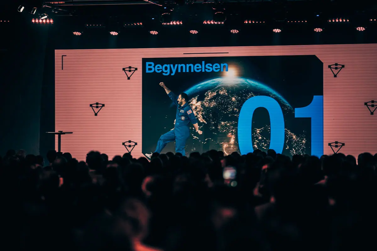 Fullsatt sal foran storskjerm med «Begynnelsen 01» under Volum